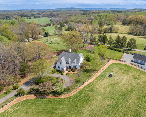 An aerial view of a charming white cottage-style home with a steeply pitched dark roof, nestled among vibrant autumn trees in hues of orange, yellow, and red. The home features dormer windows and a brick walkway leading to the front entrance, surrounded by a landscaped yard. In the background, open fields and rolling hills stretch towards a scenic horizon with distant mountain views, creating a tranquil and picturesque setting.
