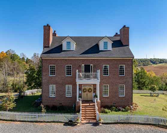 An aerial view of a stately brick manor house set within an expansive pastoral landscape. The home features a circular driveway and is accompanied by a white outbuilding with a dark roof. Surrounding the property are open fields bordered by wooden fencing, with a few horses grazing in the distance. The scene is framed by rolling hills, patches of trees, and a clear blue sky, creating a serene and picturesque countryside setting.