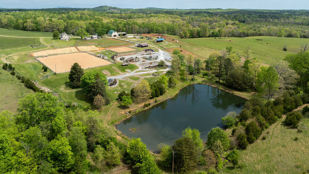 Aerial view of a Virginia horse farm featuring riding arenas, barns, fenced pastures, and a scenic pond surrounded by lush green trees and rolling countryside.