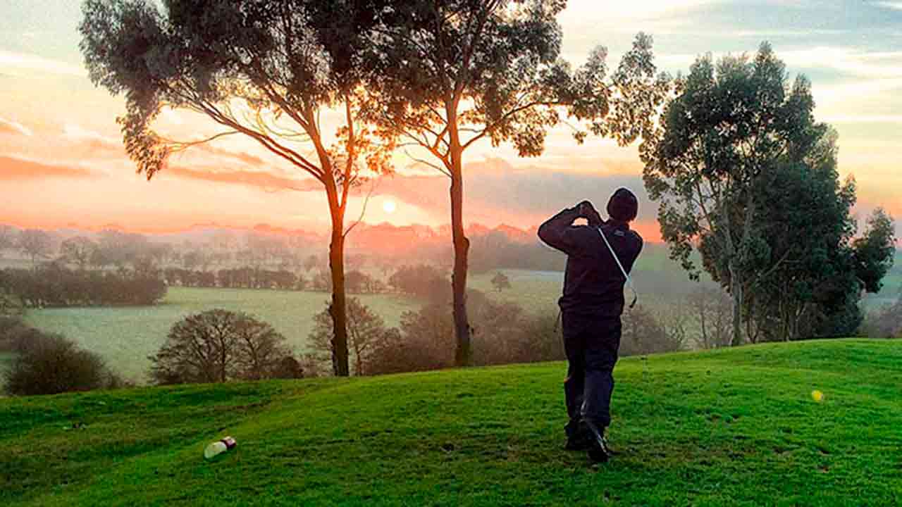 A golfer takes a swing at sunrise on a lush, green fairway surrounded by tall trees and misty countryside in Central Virginia.